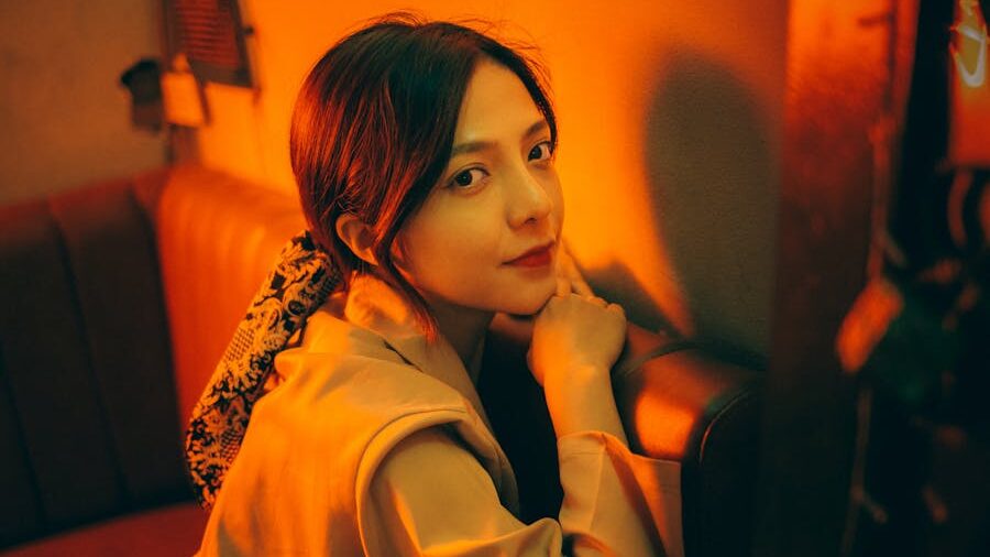 Ladyboy with dark hair and patterned scarf seated in a booth, looking at the camera under soft orange and yellow lighting.