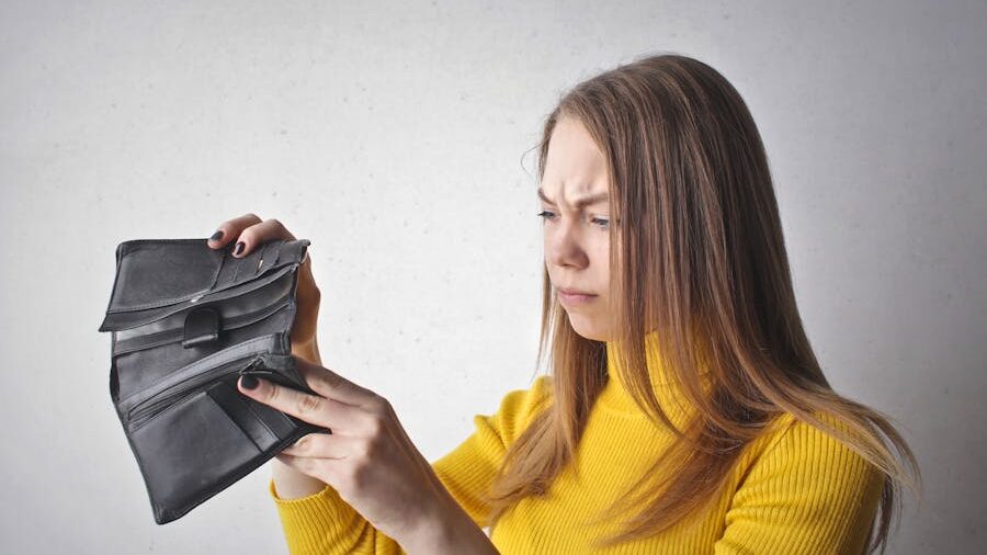 Ladyboy in a yellow sweater looking into an open, empty black wallet with a concerned expression against a light background.