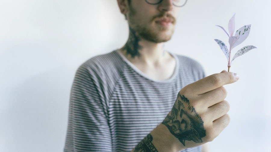 A tattooed person in a striped shirt gently holds a paper-crafted plant with pale lavender leaves against a white background.