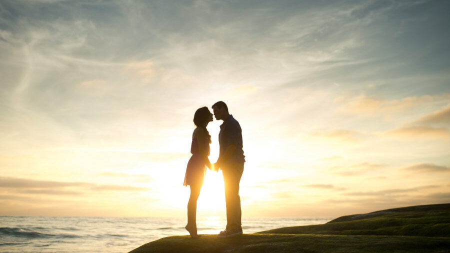 Silhouette of a couple kissing at sunset on a coastal cliff, with a glowing horizon over the ocean in the background.