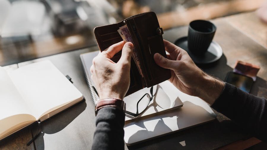 Person holding an open leather wallet with banknotes, seated at a table with a notebook, glasses, and a cup of coffee.
