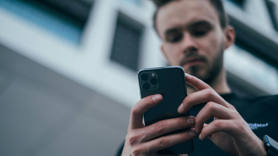 A man is holding and using a smartphone with both hands, standing outside in front of a modern glass and concrete building.