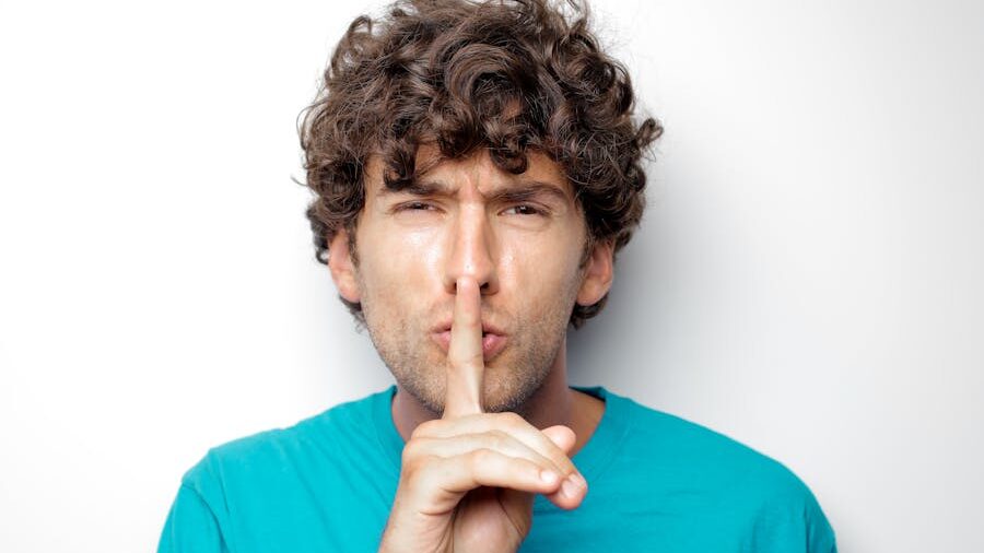 Man with curly hair holding index finger to lips in a ‘shh’ gesture, looking serious, against white background.