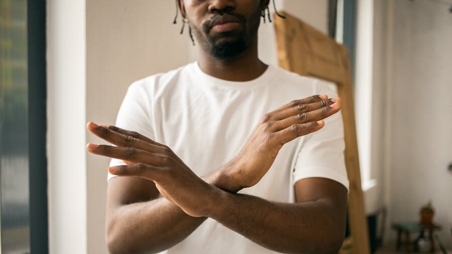 Man in white t-shirt standing indoors with arms crossed in an X to symbolize refusal or avoidance.