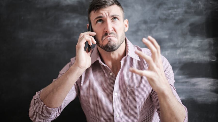 Frustrated man talking on the phone, frowning and gesturing with his hand in front of a dark chalkboard background.