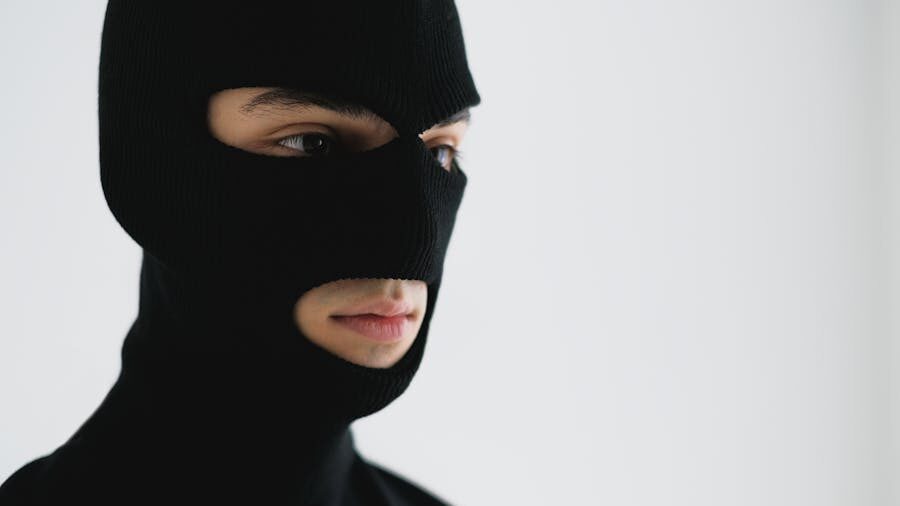 Close-up of a person in a black ski mask, facing sideways in a minimal white background.