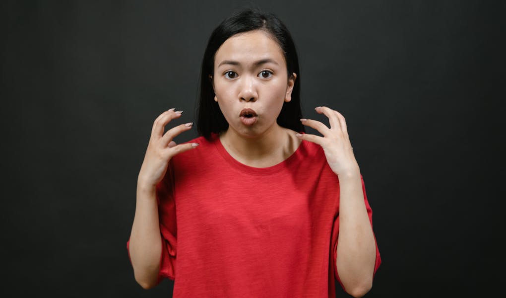 Woman in red shirt with surprised and frustrated expression, hands raised and mouth open, on black background.