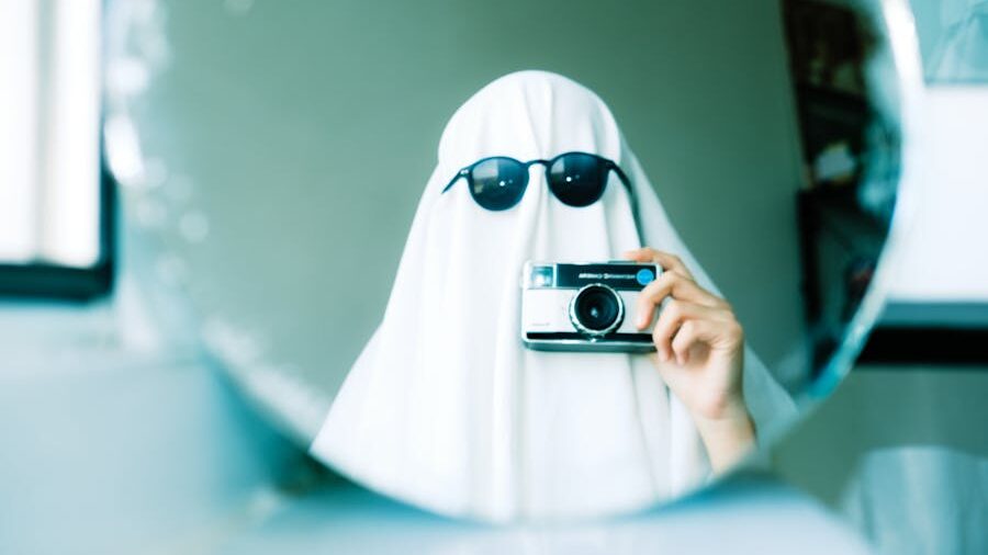 Person wearing a white sheet like a ghost with sunglasses, taking a mirror selfie using a vintage camera.