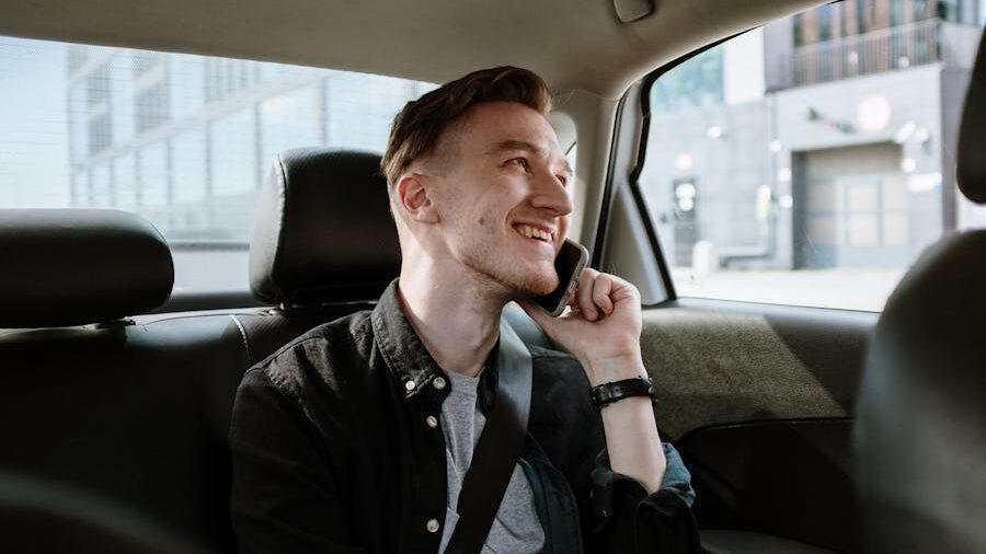 Smiling man talking on a smartphone while sitting in the back seat of a car, wearing a black shirt and seatbelt.