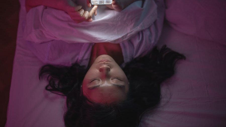 Woman lying in bed at night, holding phone above her face, screen glowing, eyes closed.
