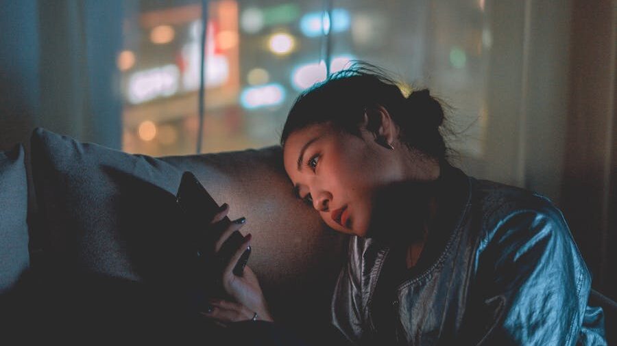 Woman in dim room staring at her phone, looking thoughtful, city lights blurred outside window.
