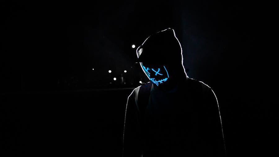 A person in a hoodie stands in the dark wearing a glowing blue mask with X eyes and stitched mouth.