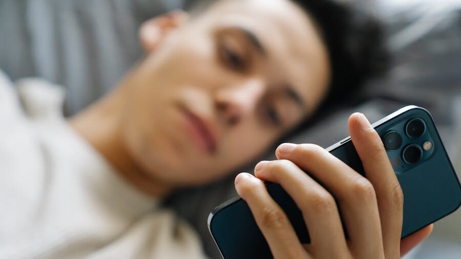Young man lying on a bed holding a smartphone in his hand, looking at the screen with a relaxed expression.