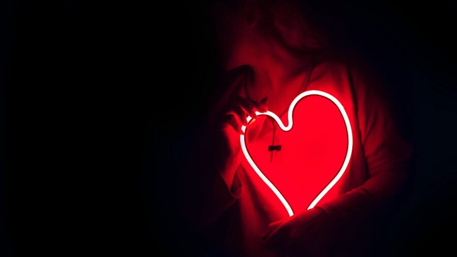 Person holding a bright red neon heart sign in a dark room, wearing a loose shirt with a necklace barely visible.