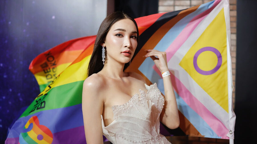 Miss Tiffany Universe 2025 contestant in white dress posing with Pride and intersex flags at Bangkok Pride Party.