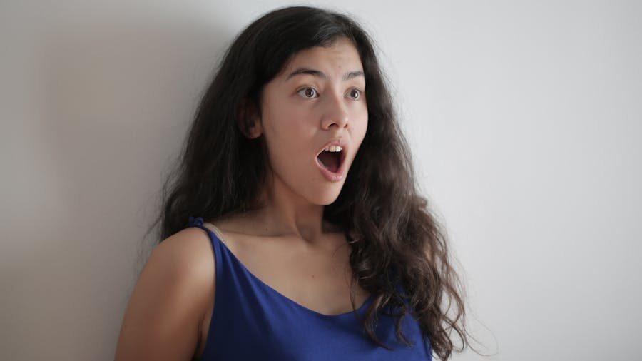Surprised young woman with long dark hair and open mouth, wearing a blue dress, standing against a plain white wall.