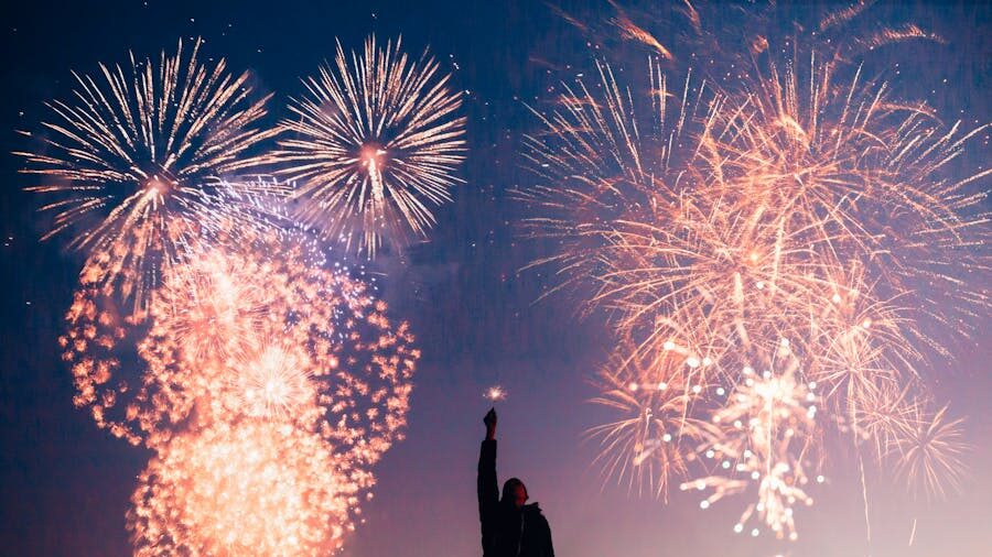 Fireworks burst above a silhouetted figure standing under the evening sky with trees in the distance.