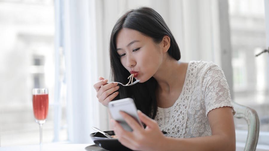 Woman with long black hair eating pasta while checking her phone, seated by a window with a glass of rosé nearby.