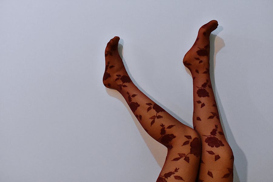 Person lying down with legs raised against a plain wall, wearing sheer black tights with a red floral pattern.