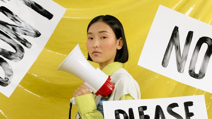 Individual in front of a yellow wall surrounded by protest signs, holding a megaphone and facing forward with determination.