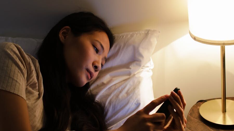 Woman lying in bed at night looking at her smartphone, illuminated by a bedside lamp in a dimly lit room.