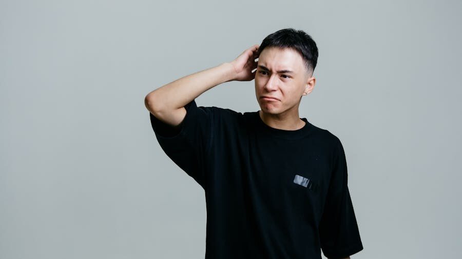 Individual standing with a puzzled look, hand behind head, wearing a black shirt in a studio setting.