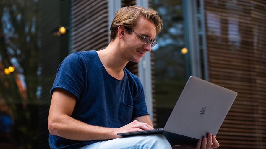 Young man sitting outside using a silver laptop, smiling slightly while looking at the screen.