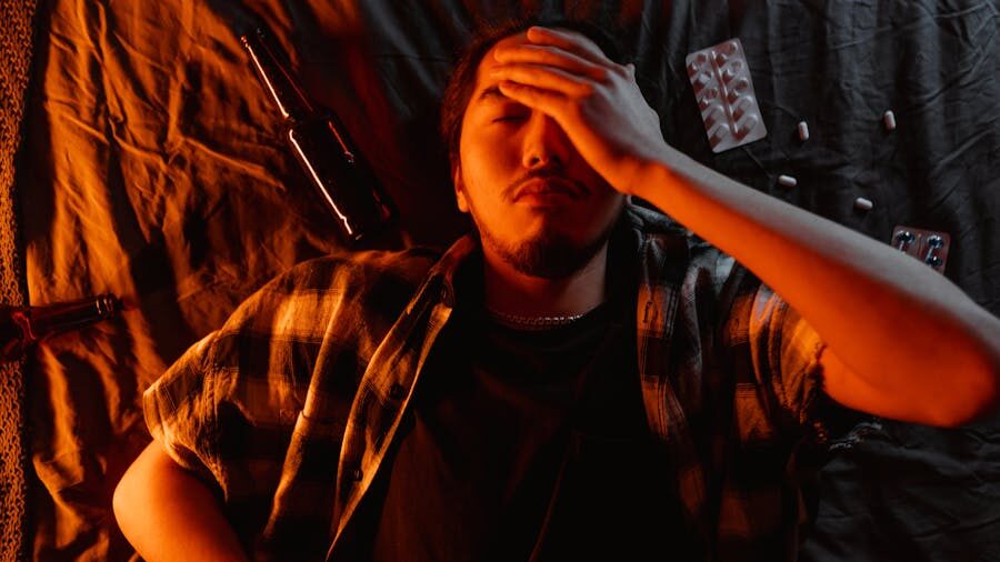 Man lying on a bed with his hand on his forehead, surrounded by pill packs and bottles in a dim orange light.