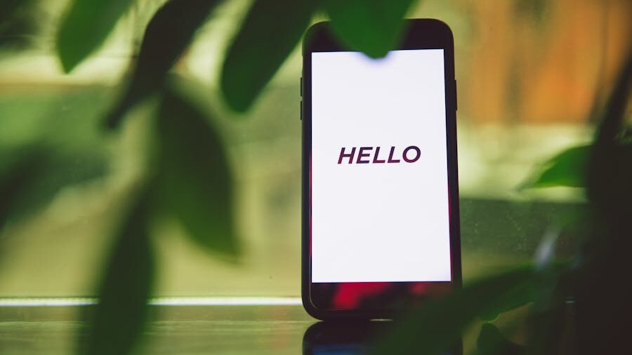 Smartphone standing upright with a white screen displaying the word “HELLO” in black text, framed by green plant leaves.