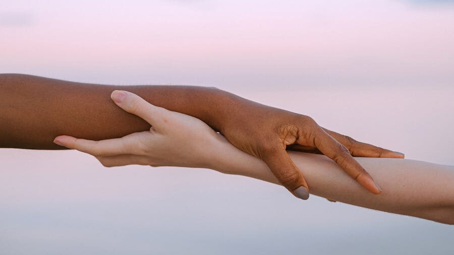 One hand gently holding another’s forearm, symbolizing unity and compassion with sky in background.