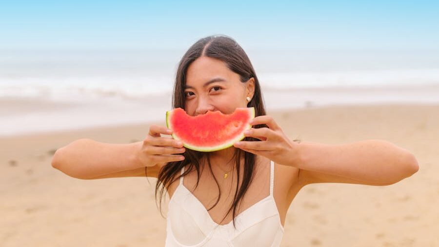 Woman at the beach holding a piece of watermelon, smiling with eyes visible against a bright seaside backdrop.