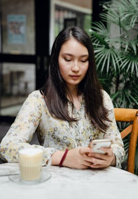 Person sitting at a café table, looking at a smartphone, with a cup of coffee in front and plants in the background.