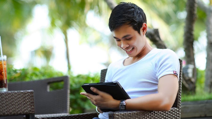 Person sitting outdoors on a chair, smiling while holding and looking at a tablet, surrounded by greenery.