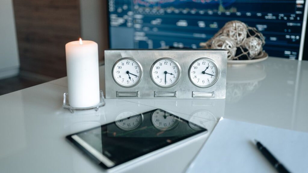 Three clocks displaying different times on a desk beside a candle, tablet, and notebook in an office setting.