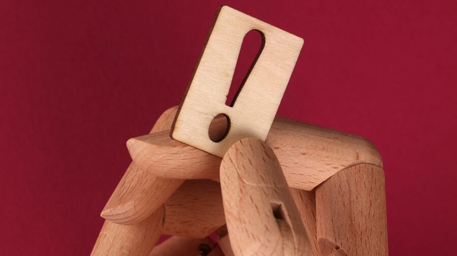 Wooden articulated mannequin holding a rectangular sign with an exclamation mark against a red background.