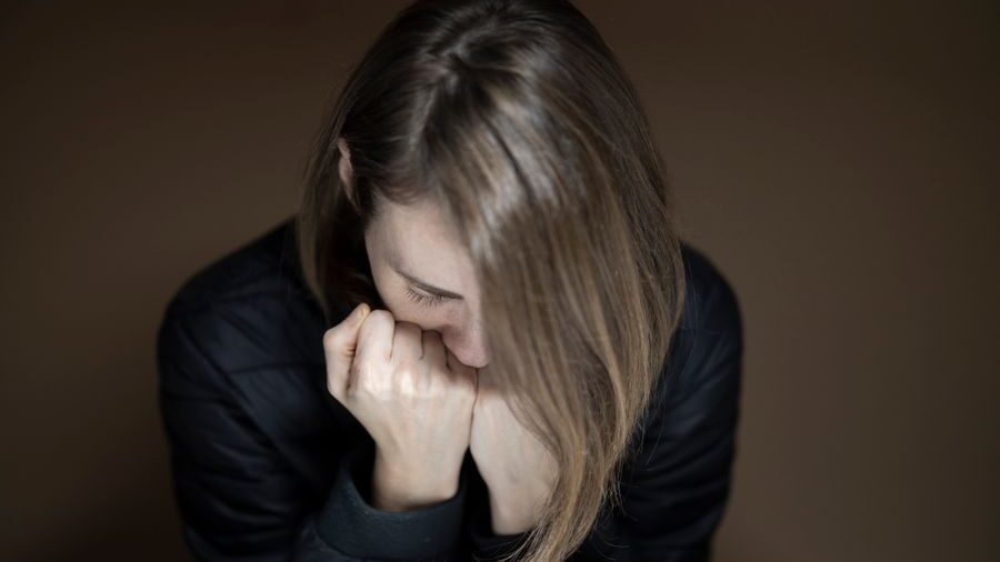 Woman with long hair looking downward while resting her face against her hands, seated against a dark background.