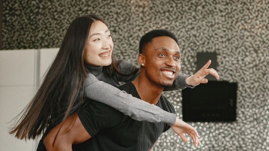 Smiling woman gives a piggyback ride to a smiling man indoors while both look toward the same direction.