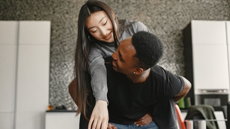 Smiling couple indoors as a woman rides on a man’s back, sharing a playful moment in a modern room.