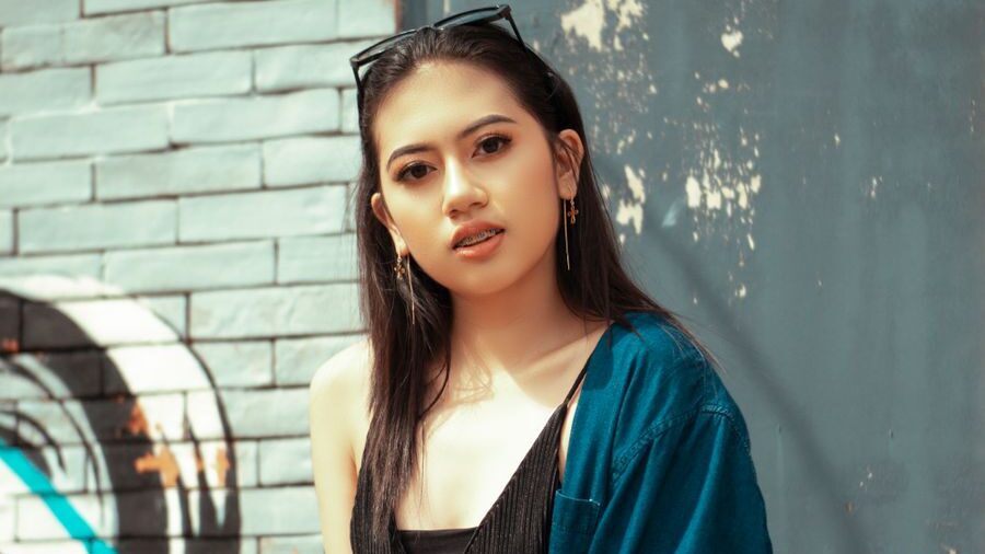 Young woman with long dark hair posing outdoors beside a gray brick wall, wearing earrings and a blue cardigan.