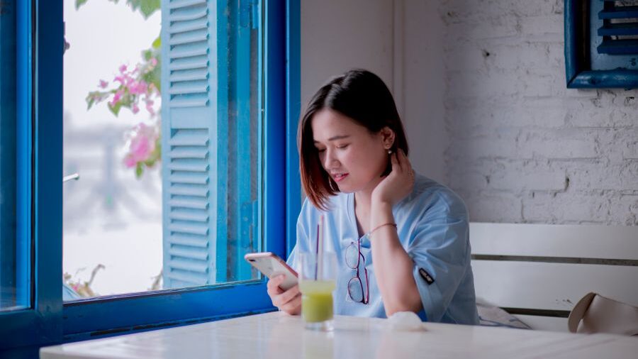Woman seated at a table near a blue-framed window, looking at her smartphone while natural light fills the room.