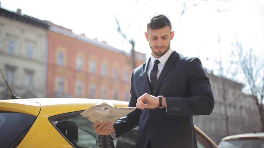 Man in a dark suit standing beside a yellow car, checking his wristwatch while holding papers outdoors.