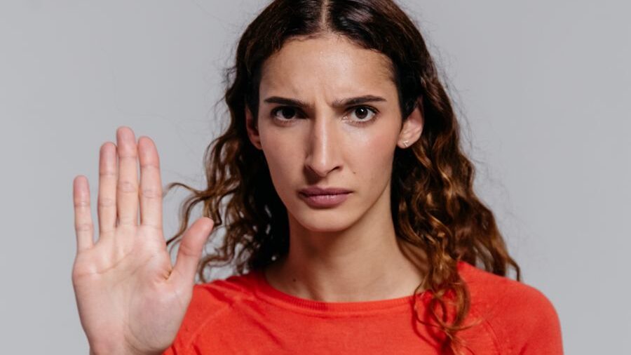Woman with curly hair wearing a red top raises her hand toward the camera with a serious expression on a plain background.