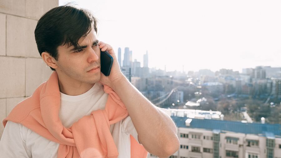Young man wearing a white shirt and pink sweater speaks on a smartphone while standing outdoors with a city skyline behind him.