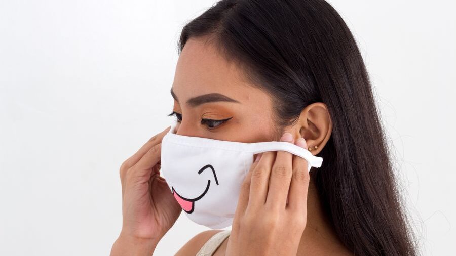 Woman with long dark hair putting on a white face mask decorated with a smiling mouth and pink tongue.