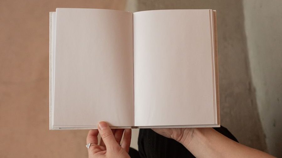 Person holding an open book with completely blank pages in front of their face, leaving their identity hidden from view.