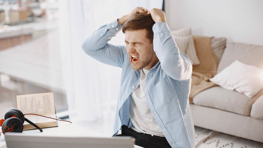 Frustrated man sitting at a desk with his eyes closed, shouting and pulling his hair while looking at a laptop.