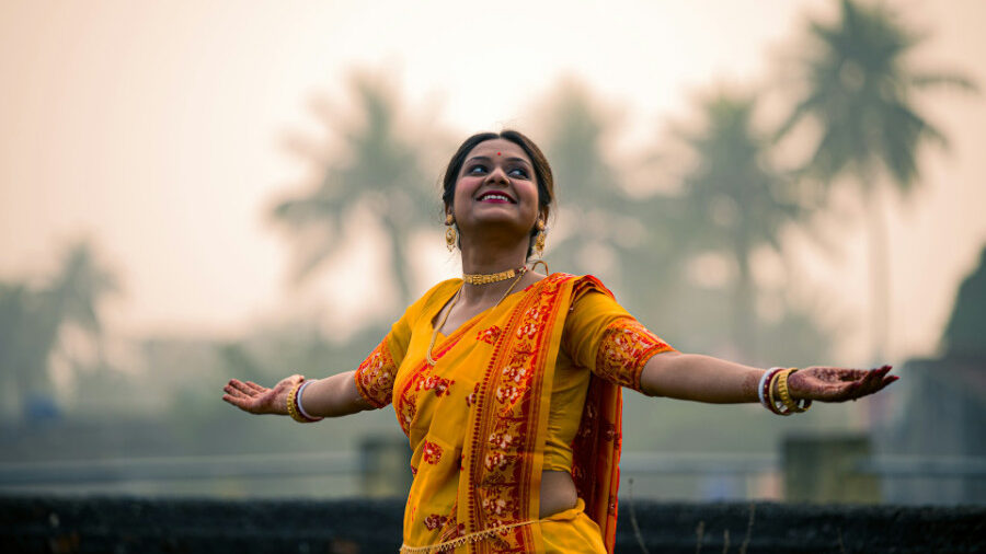 Une Hijra vêtue d'un sari jaune brodé de rouge tend les bras en souriant sur un fond d'arbres dans la lumière du petit matin.