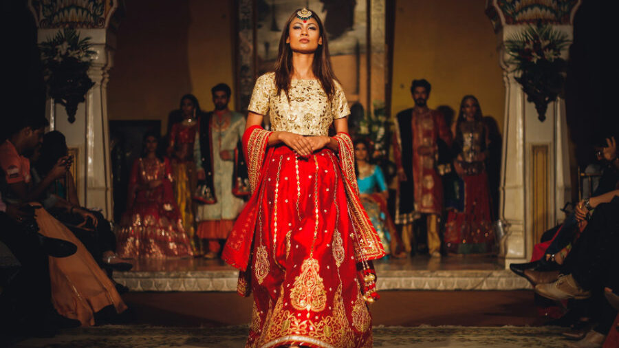 Hijra vêtue d'une tenue élaborée rouge et or sur un podium de mode, avec des spectateurs à l'arrière-plan.