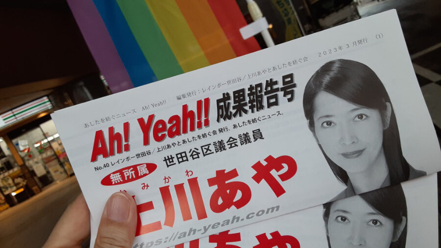 Gros plan sur des documents de campagne de Kamikawa Aya, avec son portrait et le texte 'Ah! Yeah!!' en arrière-plan, un drapeau arc-en-ciel. Prise avant la reprise de ses discours publics à Tokyo.