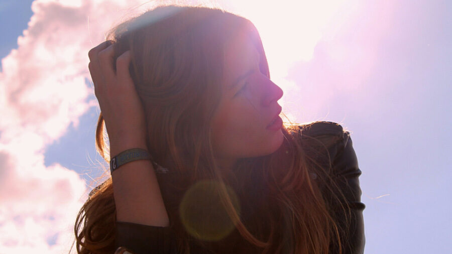 Portrait à contre-jour d'une femme se touchant ses cheveux, la lumière du soleil et les nuages créant une atmosphère douce.